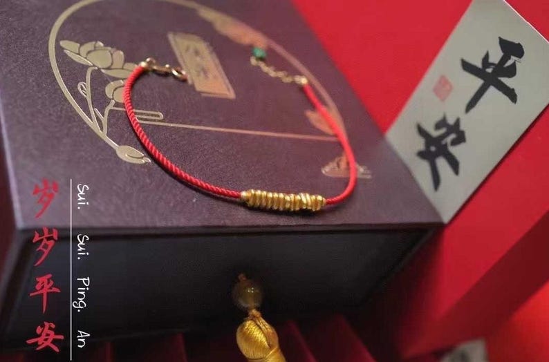 May include: A red string bracelet with a gold charm. The bracelet is on a dark brown box with Chinese characters. The box is labeled "Sui. Sui. Ping. An."