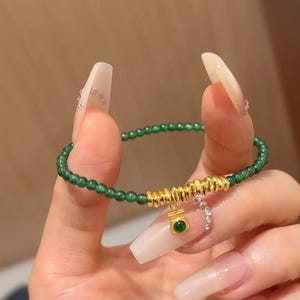 May include: A green jade bead bracelet with a gold charm featuring a green jade stone and Chinese characters. The charm is attached to a gold chain.