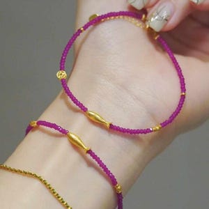 Könnte beinhalten: Drei Armbänder mit rosa Perlen und goldenen Akzenten. Das obere Armband hat einen kleinen goldenen Kreisanhänger und zwei goldene Fischanhänger. Das mittlere Armband hat zwei goldene Fischanhänger. Das untere Armband ist eine dünne goldene Kette.