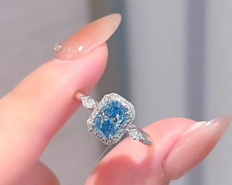 Könnte beinhalten: Ein silberner Ring mit einem rechteckigen, hellblauen Edelstein, umgeben von kleineren, klaren Steinen. Zusätzliche klare Steine ​​sind auf dem Band auf beiden Seiten des Hauptsteins angebracht. Der Ring wird von einem Finger gehalten.