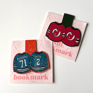 May include: Two hockey jersey bookmarks. One pair is red with white accents, the other is teal with white accents. Each bookmark has a pink background with hockey-themed illustrations and the word "bookmark".
