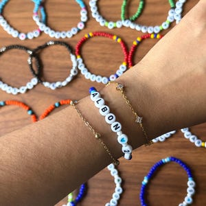 May include: A collection of colorful beaded bracelets, some spelling out the name "ALBON" with a heart symbol and the number "2". Other bracelets feature various color combinations, including blue, green, red, and orange, with white letter beads.