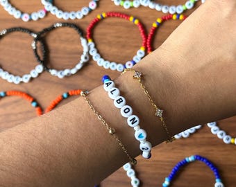 May include: A collection of colorful beaded bracelets, some spelling out the name "ALBON" with a heart symbol and the number "2". Other bracelets feature various color combinations, including blue, green, red, and orange, with white letter beads.