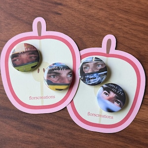 May include: Four circular pin badges featuring close-up images of eyes, each with the text "APPLE OF MY EYE." The badges are on pink apple-shaped cards with the text "florscreations."