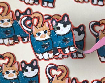 May include: A collection of die-cut stickers featuring two cats standing side-by-side, wearing teal jerseys with a shark logo. The cats have tails forming a heart shape above them. The stickers have a holographic star pattern finish on the jerseys.