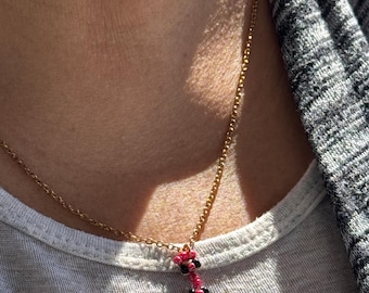 May include: A gold chain necklace with a small pendant made of pink and black beads. The pendant is shaped like a small figure.