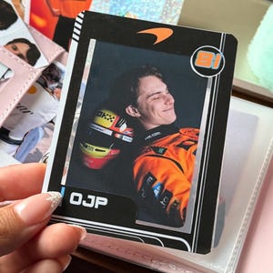 May include: A trading card featuring a smiling person in an orange racing suit and helmet. The card has a black border with the McLaren logo and the initials "OJP". The number "81" is in a circle on the upper right corner.