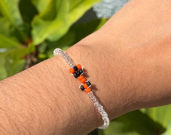 May include: A beaded bracelet featuring a series of clear, translucent beads interspersed with a central pattern of alternating small black and bright orange beads. The bracelet is constructed on a thin, flexible cord and sits on a wrist.