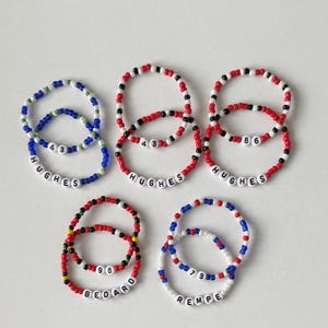 May include: Six beaded bracelets with red, white and blue beads. The bracelets have names and numbers on them, including "Hughes 43", "Hughes 86", "Bedard 98", and "Rempe 73".