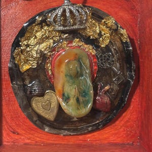May include: A mixed media artwork with a central, colourful, abstract shape within a circular frame. The frame includes a small silver crown, gold leaf accents, and miniature charms like a heart, key, and beehive, all set against a red background.