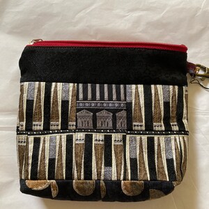 May include: A black zippered pouch with a red zipper. The pouch has a patterned fabric with a design of columns and geometric shapes in shades of brown, black, and white.