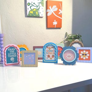 Op de afbeelding: Een verzameling kleurrijke decoratieve frames met verschillende ontwerpen en tekst, waaronder "Enjoy the Little Things" en "Good Vibes". De frames zijn gerangschikt op een wit oppervlak, met kunstwerken aan de muur op de achtergrond.
