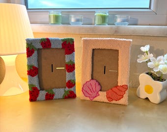 May include: Two framed pictures with different designs. The first frame is blue and white with a strawberry pattern. The second frame is pink with a seashell design.