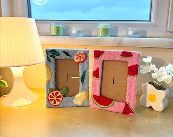 May include: Two framed pictures with colorful embroidery. One frame has a blue background with lemons, oranges, and leaves. The other frame has a pink background with watermelons.