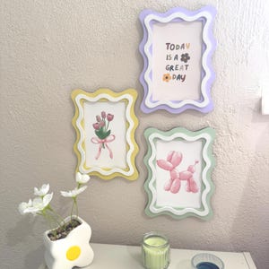May include: Three decorative picture frames in yellow, purple, and green with wavy edges. The frames display illustrations of flowers, a balloon dog, and the text "TODAY IS A GREAT DAY". A small vase with white flowers and a candle are also visible.