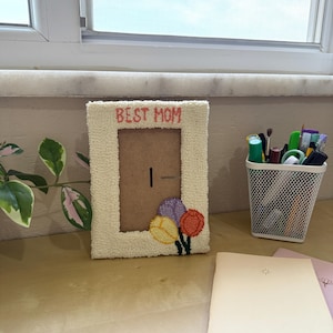 May include: A cream-coloured photo frame with the text "BEST MOM" in pink. The frame has a textured, fluffy border and features a bouquet of embroidered flowers in yellow, purple, and orange. The frame is on a wooden surface.