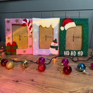 May include: Three Christmas-themed picture frames with different designs. The first frame is pink with gold stars, a candy cane, and a wrapped present. The second frame is light blue with a white snowy roof and a snowman. The third frame is green with a red Santa hat and the text "HO HO HO".