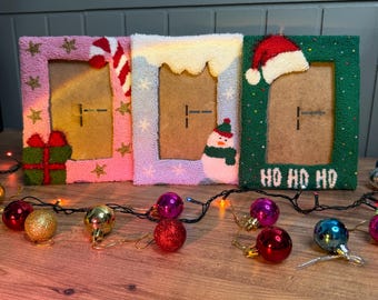 May include: Three Christmas-themed picture frames with different designs. The first frame is pink with gold stars, a candy cane, and a wrapped present. The second frame is light blue with a white snowy roof and a snowman. The third frame is green with a red Santa hat and the text "HO HO HO".