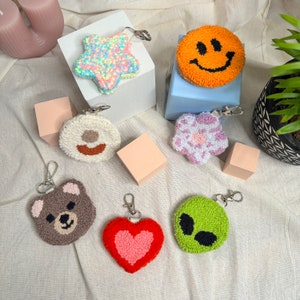 May include: A set of 10 colourful, hand-stitched keychains featuring various designs, including a star, a smiley face, a flower, a bear, a heart, and an alien. The keychains are made with a fluffy, yarn-like material and are attached to silver key rings.