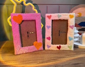 May include: Two handmade picture frames with a heart design. The frame on the left is pink with orange hearts and the word "love" written on the bottom. The frame on the right is white with multi-colored hearts.