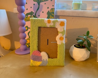 May include: A green picture frame with a white daisy design and a colorful Easter egg design. The frame is made of yarn and has a brown cardboard backing.