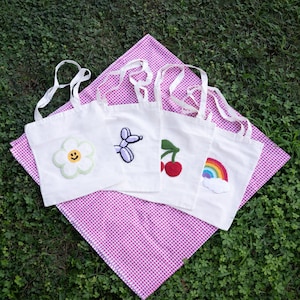 Op de afbeelding: Vier witte tote bags met verschillende geborduurde ontwerpen. De ontwerpen omvatten een bloem, een ballondier, een kers en een regenboog. De tassen worden getoond op een roze en witte gingham tafelkleed.