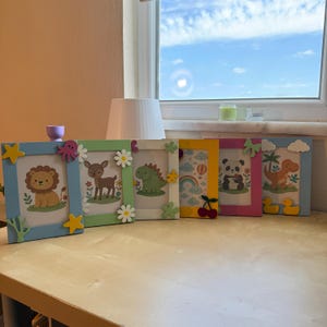 Può includere: Una collezione di sei cornici colorate con disegni di animali dei cartoni animati. Ogni cornice presenta un animale diverso, tra cui un leone, un cervo, un dinosauro, un panda e delle anatre. Le cornici sono decorate con stelle, fiori e fiocchi.