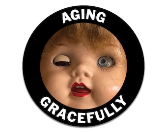 May include: A circular black sticker with the text "AGING GRACEFULLY" in white. The image shows a close-up of a vintage doll's face, with one eye closed and red lipstick, within the sticker's border.