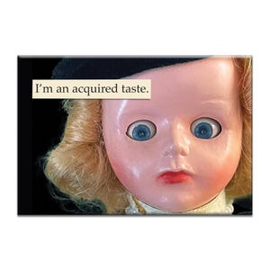 Pode incluir: Uma boneca vintage com olhos azuis e cabelo loiro, usando um chapéu preto e uma gola branca. O texto "I'm an acquired taste" é exibido em um rótulo retangular acima do rosto da boneca. A pele da boneca é rosa pálido.