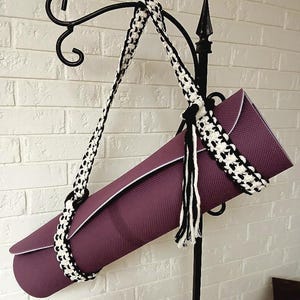 以下が含まれることがあります： 白のトリムが付いた、丸められた紫色のヨガマット。白と黒の編み込みストラップで固定されています。マットは白いレンガの壁を背景に、黒い金属製のスタンドから吊り下げられています。タッセルがストラップからぶら下がっています。