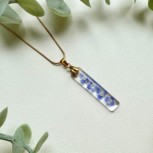 Könnte beinhalten: Eine goldene Kettenkette mit einem klaren rechteckigen Anhänger, der gepresste blaue Blumen enthält.