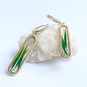 May include: Gold hoop earrings with a clear rectangular pendant featuring a pressed green leaf design.