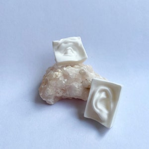 May include: Two white ceramic sculptures, one shaped like a face and the other like an ear, are displayed on a pink crystal rock.