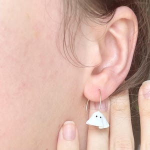 Peut inclure: Boucles d'oreilles pendantes en forme de fantôme blanc avec des yeux noirs. Les boucles d'oreilles sont en pâte polymère et sont suspendues à des crochets dorés.
