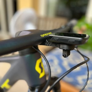 Puede incluir: Un manillar de bicicleta negro con un ordenador de ciclismo negro y gris montado en la parte superior. El ordenador tiene una pantalla que muestra datos y hay botones en el lateral. El manillar tiene un logotipo "SYMMETRIC" amarillo.