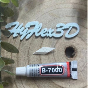 Może przedstawiać: Białe literki z brokatem tworzące napis "HyFlex 3D" na drewnianej powierzchni. Na powierzchni znajduje się również mały biały diament i tubka kleju B-7000.