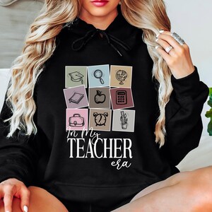May include: A black hoodie with a graphic of school supplies in colorful squares and the text "In My Teacher Era".
