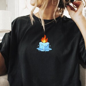 May include: Black t-shirt featuring a graphic of a blue ice cube with a flame on top. The person is sitting on a light-colored couch and holding a white mug. The person is wearing white shorts and fuzzy slippers.