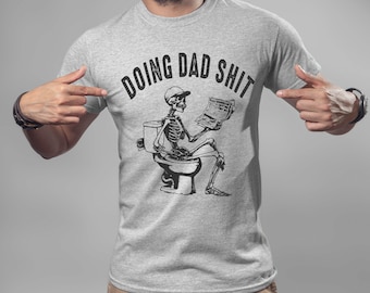 May include: A light gray t-shirt with the phrase "DOING DAD SHIT" printed across the top. Below the text is a black and white graphic of a skeleton sitting on a toilet, reading a newspaper. The shirt is being pointed at by a person.