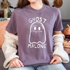 May include: A purple t-shirt with a white ghost graphic. The ghost has two oval eyes and the words "GHOST MALONE" above and below it. The ghost graphic also includes the words "Always Tired" and other small details.