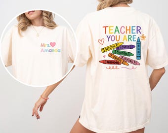 May include: Cream-colored t-shirt with the text "TEACHER YOU ARE" in rainbow colors and crayon graphics. The front of the shirt has the text "Mrs. Amanda" in pastel colors with a heart symbol. A cute and colorful design for teachers.