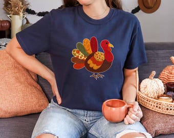 May include: Navy blue t-shirt featuring a colorful turkey design. The turkey has a red head, yellow beak, and a patchwork of autumn-toned feathers. The shirt is a comfortable, casual style, perfect for Thanksgiving or fall.