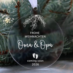 May include: Clear acrylic Christmas ornament with white text and a white ribbon for hanging. The ornament says "Frohe Weihnachten Oma & Opa" with baby footprints and "coming soon 2026". The ornament is hanging in front of a green Christmas tree.