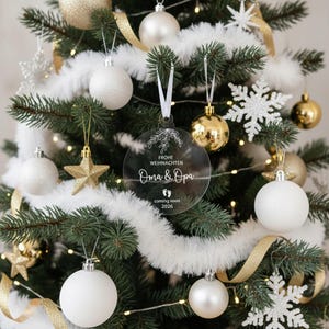 May include: A Christmas tree decorated with white, gold, and silver ornaments. A clear, round ornament with the text "Frohe Weihnachten Oma & Opa coming soon 2026" hangs in the center. The tree is adorned with white garland and gold ribbon.
