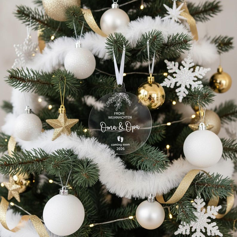 May include: A Christmas tree decorated with white, gold, and silver ornaments. A clear, round ornament with the text "Frohe Weihnachten Oma & Opa coming soon 2026" hangs in the center. The tree is adorned with white garland and gold ribbon.