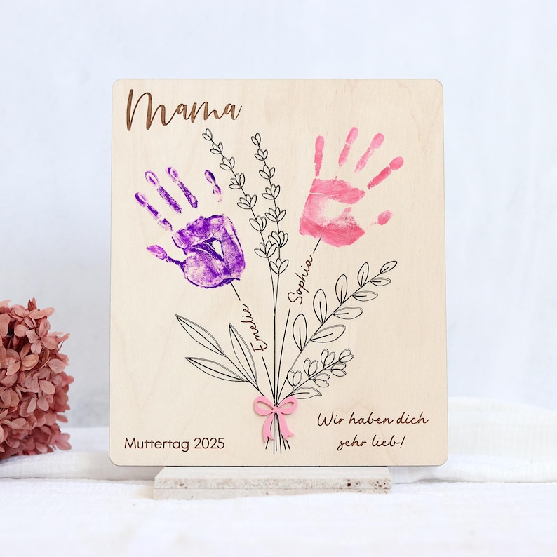 May include: Wooden sign with the word "Mama" at the top, two handprints in purple and pink, and the text "Wir haben dich sehr lieb!"  The sign also includes the text "Muttertag 2025" and the names "Sophia" and "Emelie" written next to the handprints.