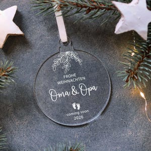 May include: Clear acrylic Christmas ornament with white text and a floral design. The ornament reads "Frohe Weihnachten Oma & Opa coming soon 2026" and includes baby footprints. A white ribbon is attached for hanging.