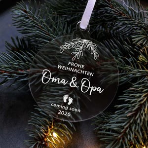 May include: A clear, round Christmas ornament with white text and a floral design. The text reads "Frohe Weihnachten Oma & Opa coming soon 2026" with baby footprints. A white ribbon is attached for hanging.