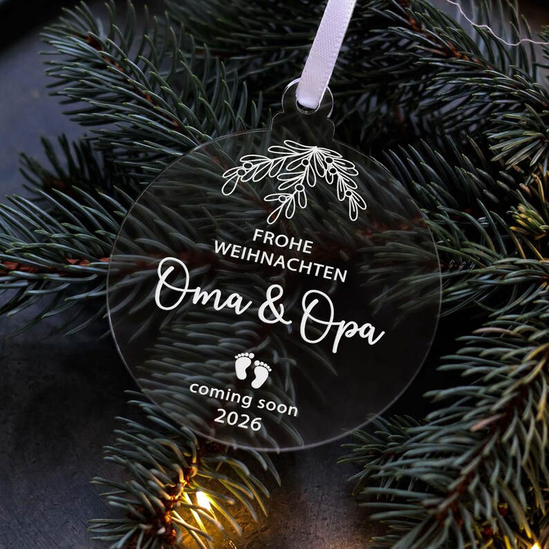 May include: A clear, round Christmas ornament with white text and a floral design. The text reads "Frohe Weihnachten Oma & Opa coming soon 2026" with baby footprints. A white ribbon is attached for hanging.
