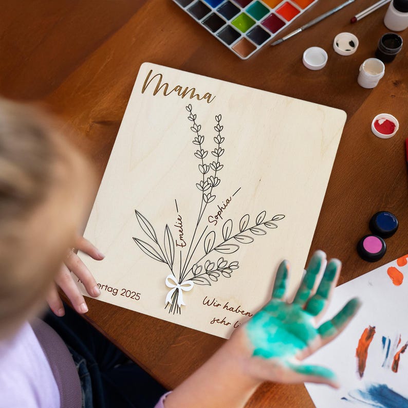 May include: A wooden sign with the word "Mama" at the top. A hand-drawn bouquet of flowers is in the center with the names "Emeliie" and "Sophia" written on the stems. The text "Wir haben sehr lieb" is at the bottom of the sign.